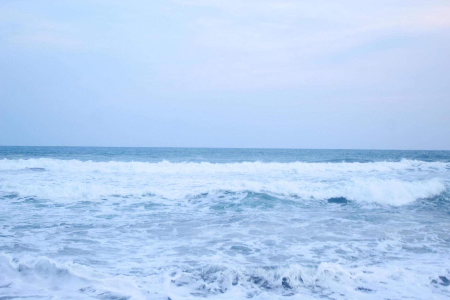 As the tide sweeps in at South Jogja Jetis Beach, Cornwall under a serene sky, waves gently roll, accompanied by ripples and bubbling foam. photo