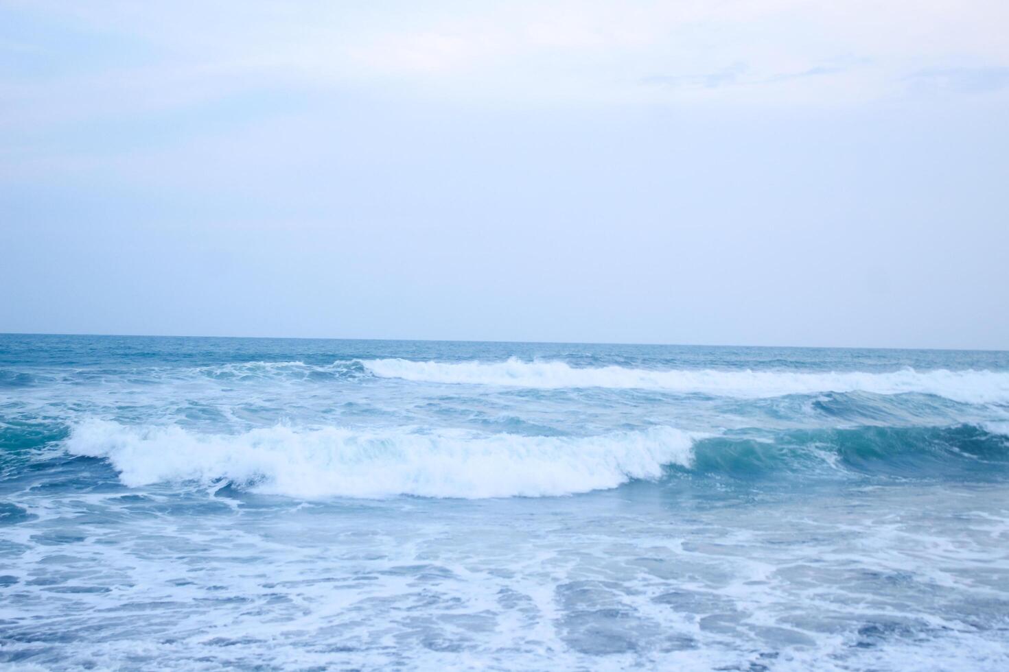 As the tide sweeps in at South Jogja Jetis Beach, Cornwall under a serene sky, waves gently roll, accompanied by ripples and bubbling foam. photo