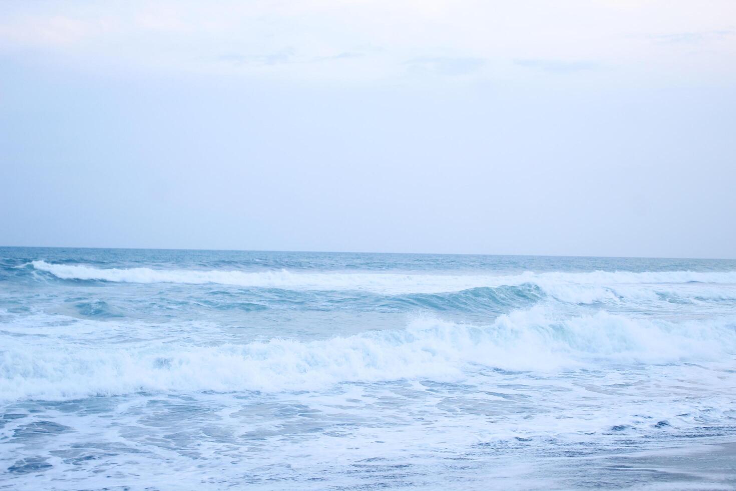 As the tide sweeps in at South Jogja Jetis Beach, Cornwall under a serene sky, waves gently roll, accompanied by ripples and bubbling foam. photo
