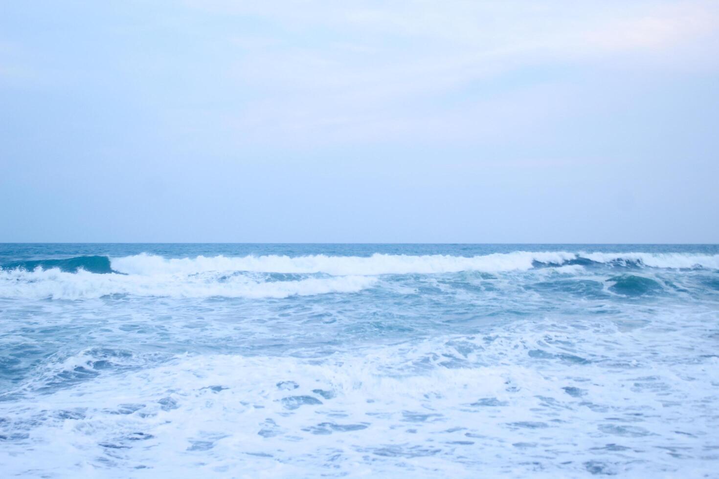 As the tide sweeps in at South Jogja Jetis Beach, Cornwall under a serene sky, waves gently roll, accompanied by ripples and bubbling foam. photo
