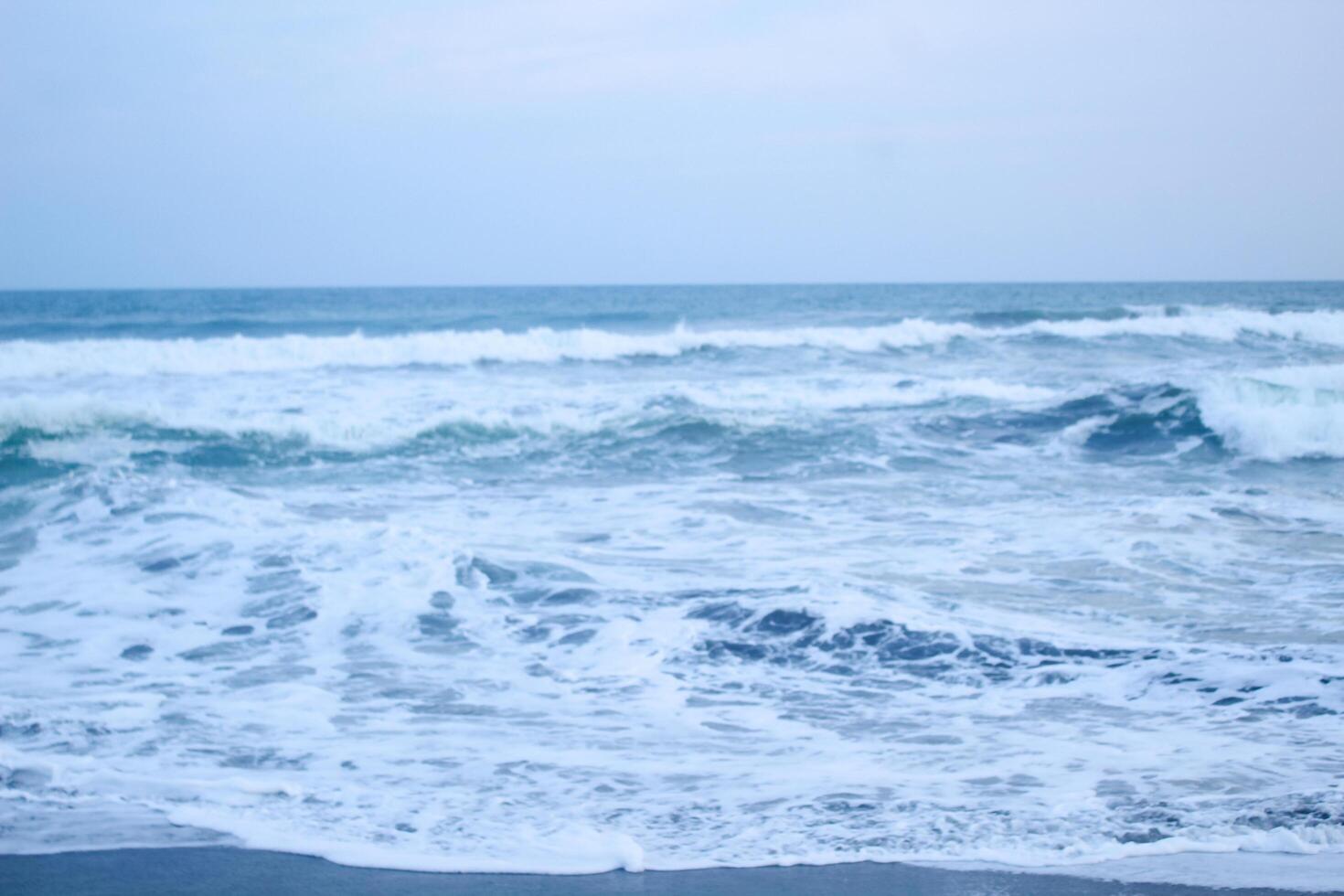 As the tide sweeps in at South Jogja Jetis Beach, Cornwall under a serene sky, waves gently roll, accompanied by ripples and bubbling foam. photo