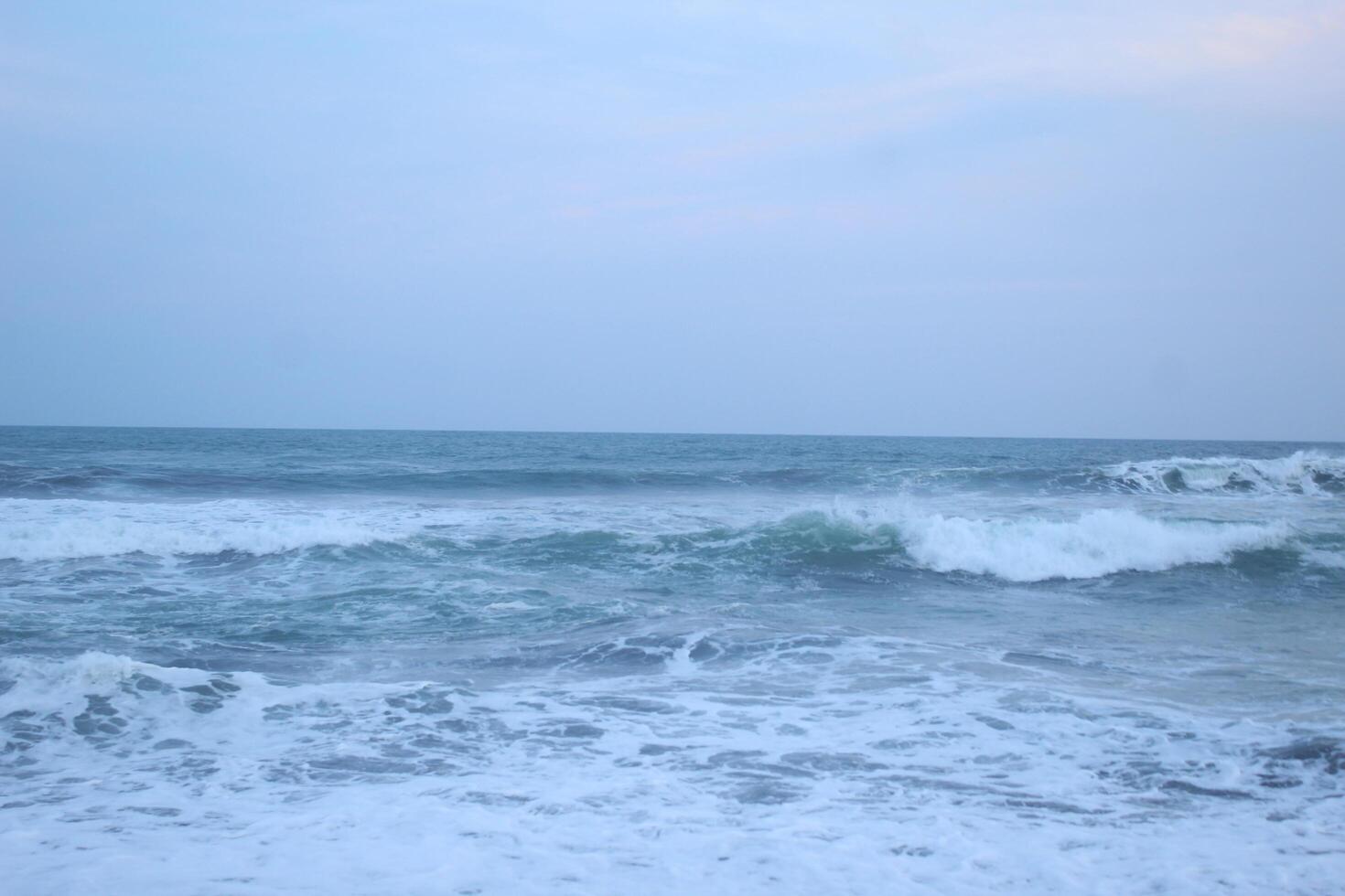 As the tide sweeps in at South Jogja Jetis Beach, Cornwall under a serene sky, waves gently roll, accompanied by ripples and bubbling foam. photo