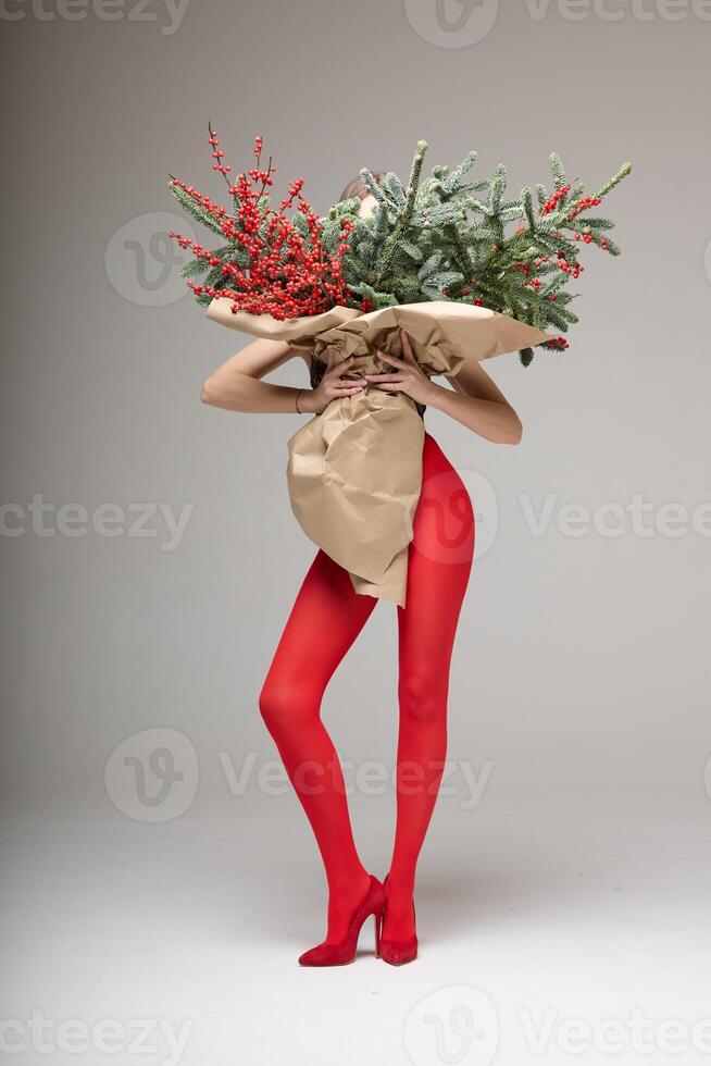 A long girl in red tights and high-heeled shoes holds a huge bouquet of fir branches and red berries wrapped in cardboard paper, she closes with a bouquet, photo studio with white background