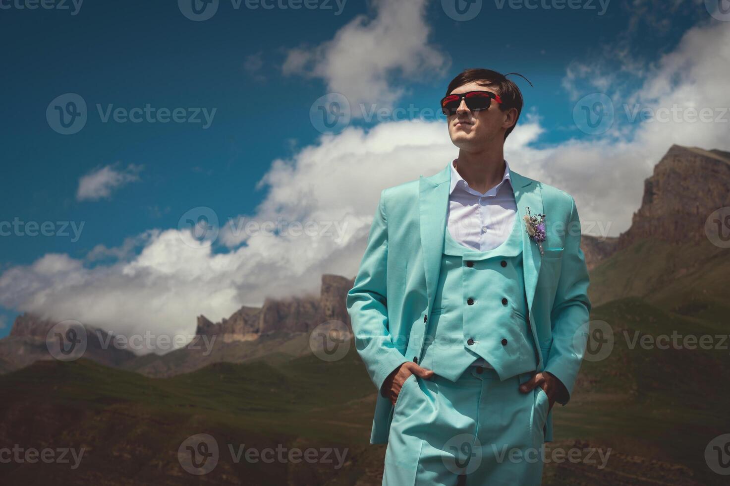Groom in a suit stands against a background of mountains and clouds in summer. A man in a turquoise three-piece suit stands confidently looking to the side photo