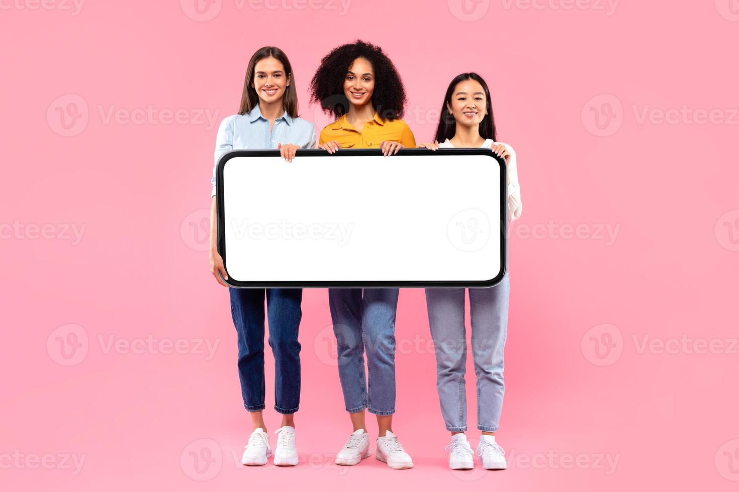 Full length shot of diverse ladies holding huge smartphone with white blank screen, offering mockup for your app or website design, pink studio background photo