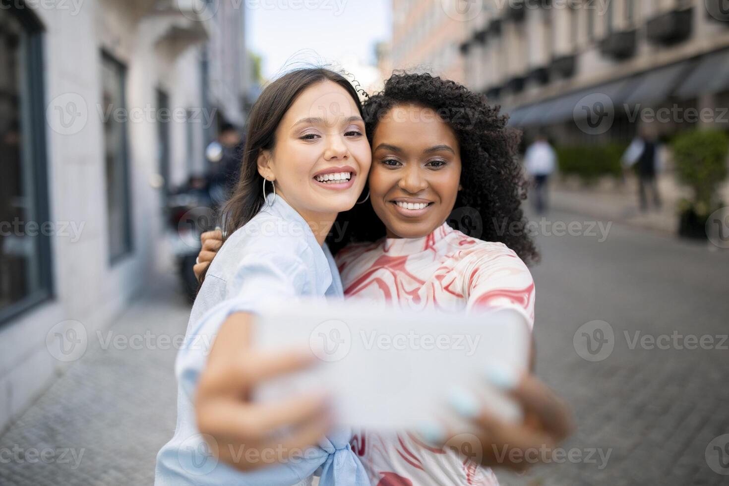 Two Beautiful Female Friends Laughing While Taking Selfie On Smartphone Outdoors, Happy Cheerful Multiethnic Besties Posing At Camera Together, Making Photos While Walking In City, Selective Focus