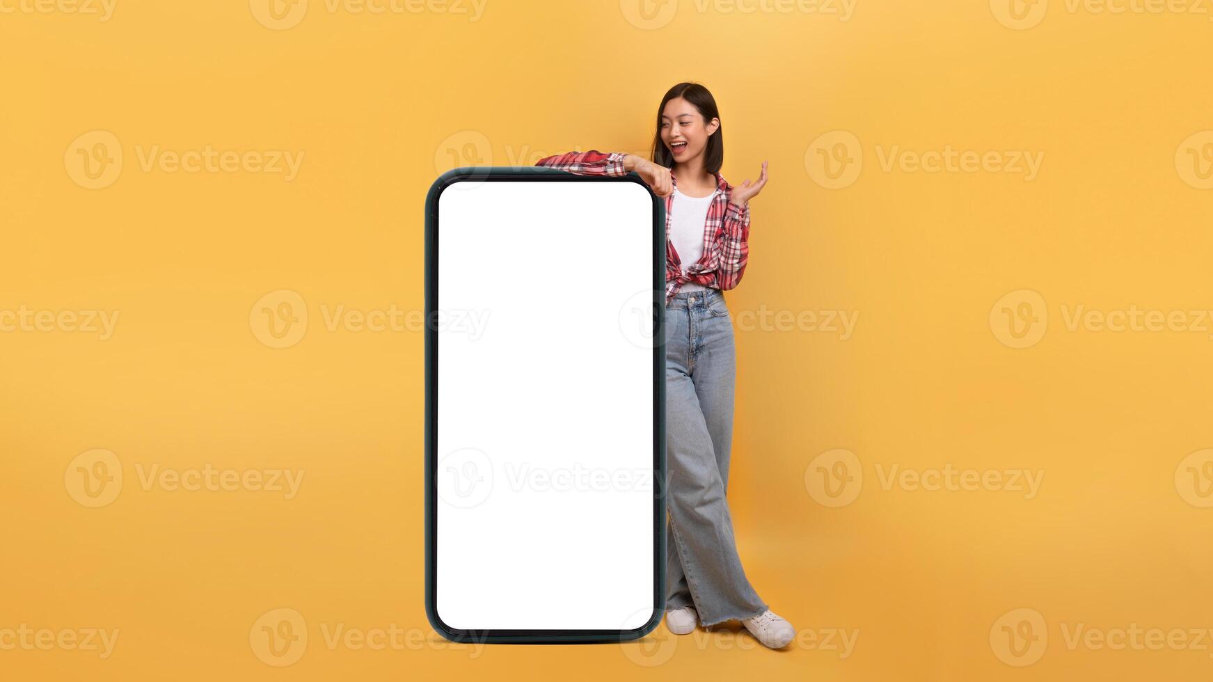 Great mobile af. Happy asian lady leaning on huge giant cellphone with blank screen and shouting wow, female standing isolated over yellow background, panorama, banner photo