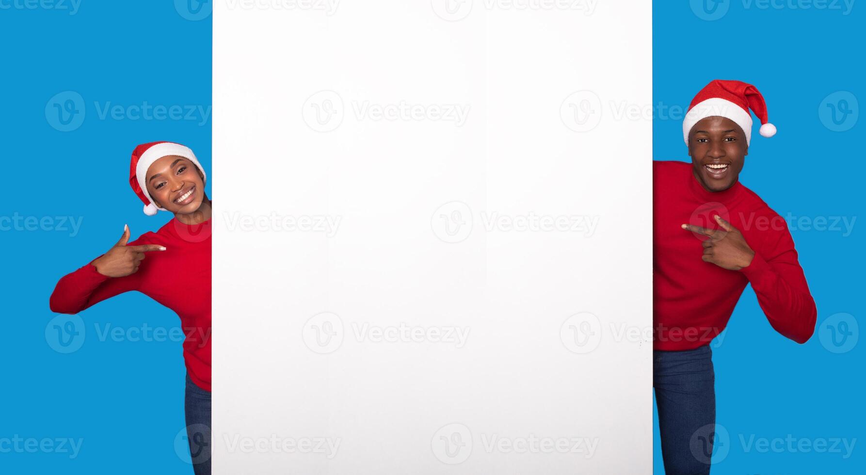 Christmas Offer. Joyful Young Black Couple In Red Santa Hats Pointing Fingers At Blank Paper Poster, Showing Free Space For Holiday Party Invitation, Posing On Blue Studio Background. Mockup photo