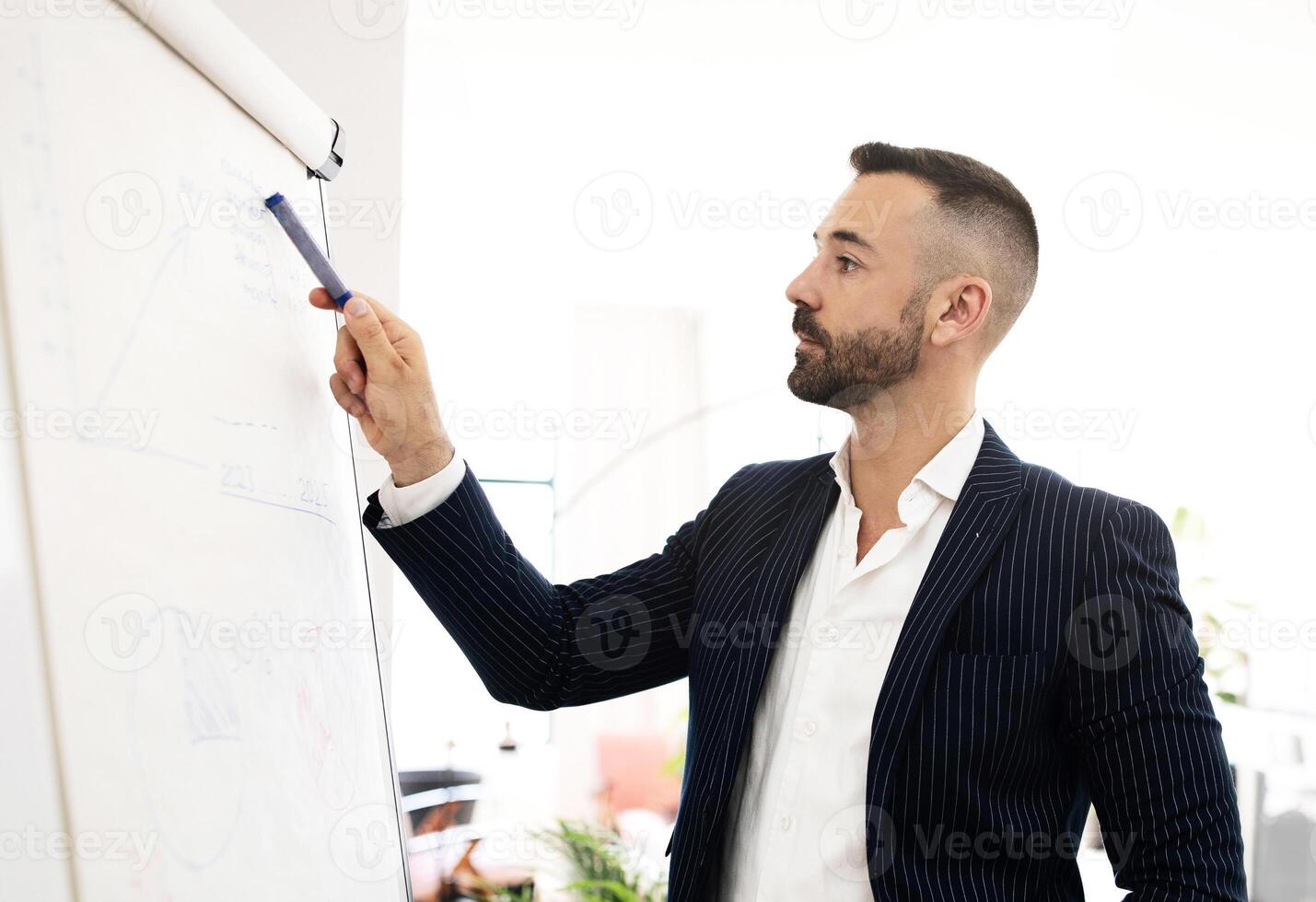 Serious confident caucasian mature man with beard in suit point at blackboard with graphs and charts in coworking office interior. Businessman, teacher, manager work, meeting and business planning photo