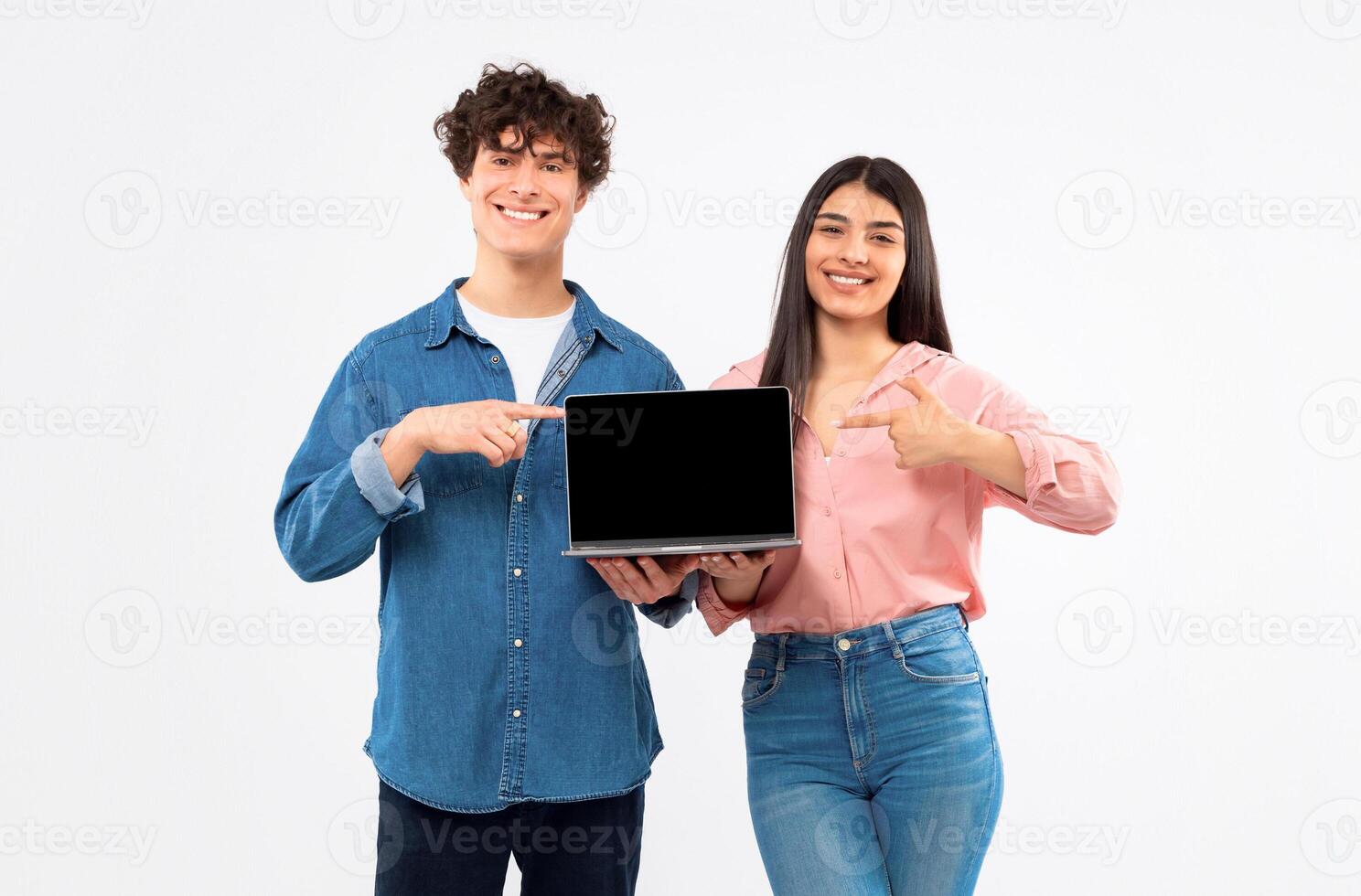 Digital Education Offer. Cheerful Young Students Couple Showing Laptop Empty Screen, Pointing Finger At Mockup For Educational Website Ad, Standing Together Over White Background, Studio Shot photo