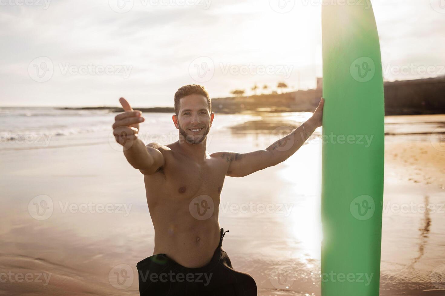 Surf With Me. Handsome Young Surfer Guy Holding Surfboard And Making Invite Gesture With Fingers At Camera, Shirtless Smiling Millennial Man Having Fun While Surfing On The Beach At Sunset photo