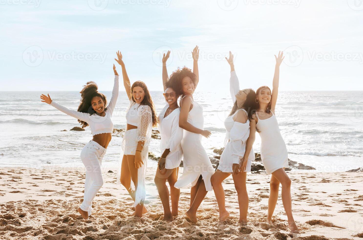 Happy multiracial ladies dancing with arms raised while celebrating hen party on the beach at sunny day, full length. Joyful young women bride's team photo