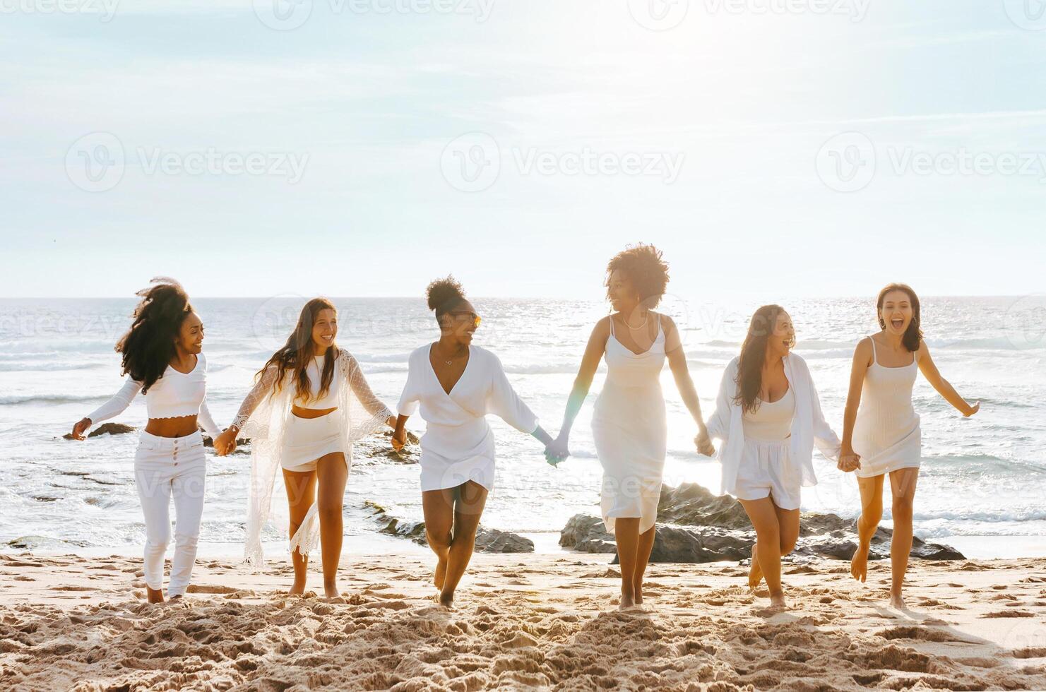 Happy diverse ladies walking on the sea shore, holding hands and laughing, having hen party on the beach, full length. Multiracial women enjoying holiday celebration photo
