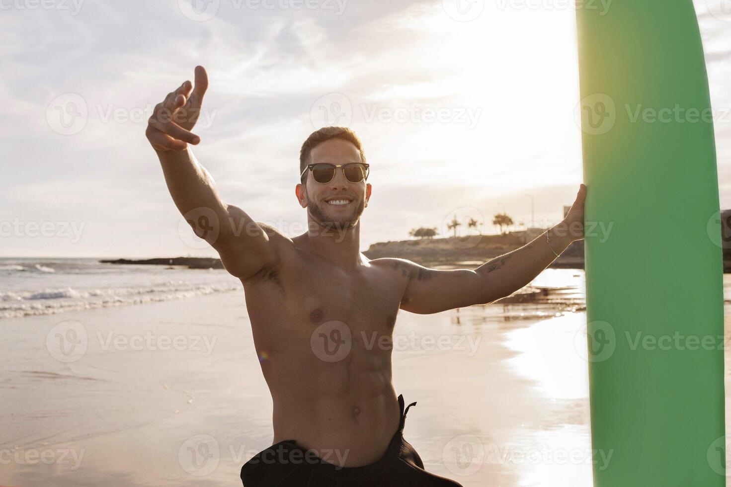 Portrait Of Young Handsome Male Surfer Posing With Surfboard On The Beach At Sunset Time, Smiling Sporty Millennial Guy In Stylish Sunglasses Pointing Fingers At Camera, Enjoying Surfing, Free Space photo