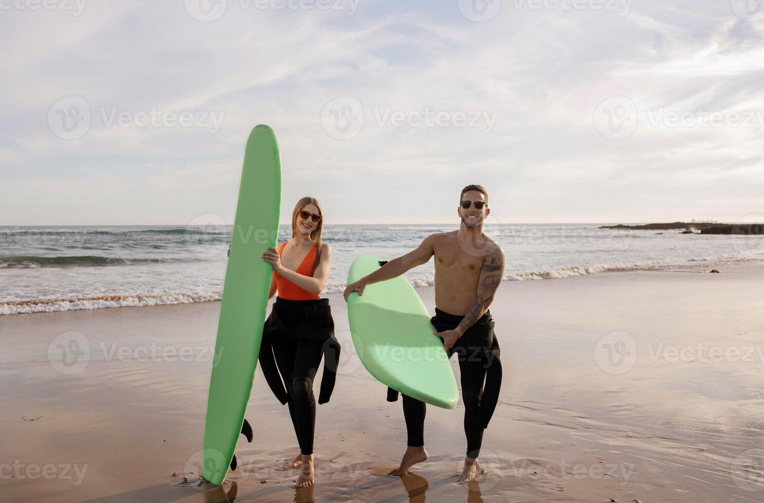 Young man and woman with surfboards posing on the beach at sunset, happy millennial surfers in stylish sunglasses having fun while surfing together, cheerful friends enjoying water sports photo