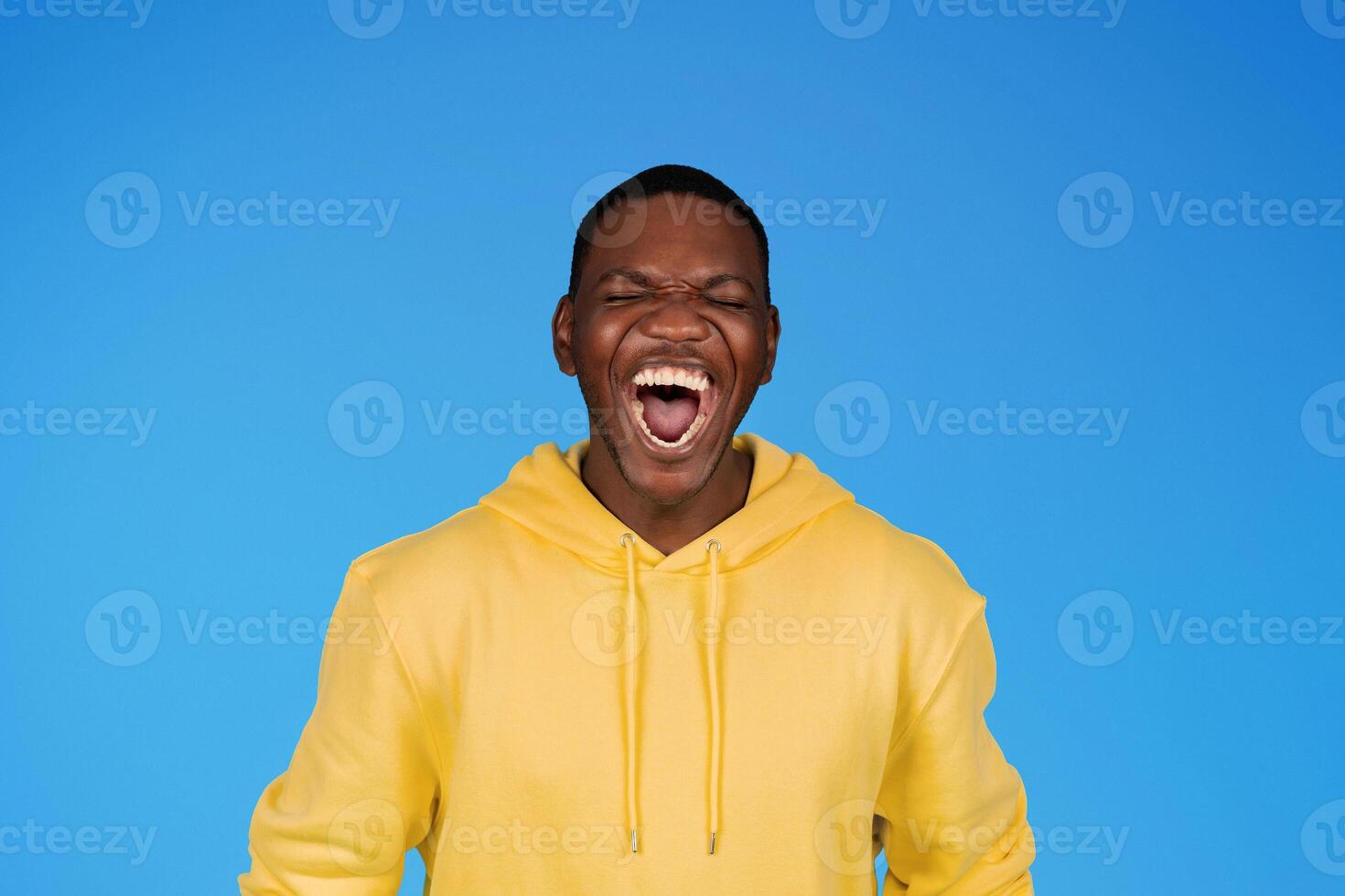 Cheerful laughing excited millennial african american guy in sweatshirt with wide mouth open, enjoy win, isolated on blue studio background. Human emotions, ad and offer, scream photo