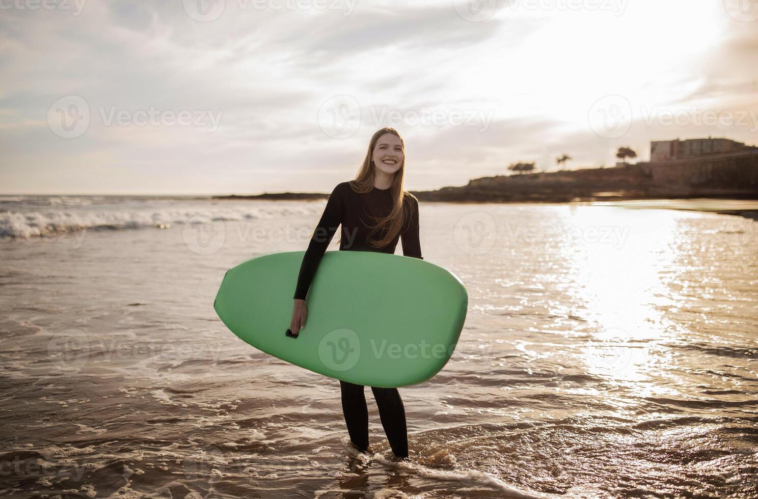 Sport Activities. Portrait Of Cheerful Young Surfer Woman Posing With Surfboard, Smiling Millennial Female Wearing Wetsuit Surfing On The Beach On Sunset, Enjoying Water Sports And Summer Vacation photo