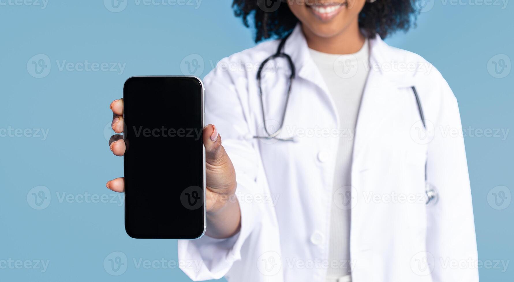 Smiling black millennial lady doctor in white coat show smartphone with empty screen, isolated on blue studio background, cropped. App for work professional recommendation, medicine and health care photo