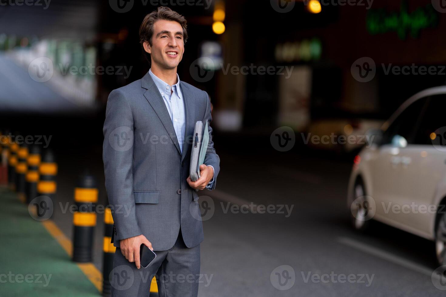 Handsome smiling millennial man wearing suit CEO manager standing at parking area, holding smartphone and laptop in his hands, waiting for his car or taxi, looking at copy space photo