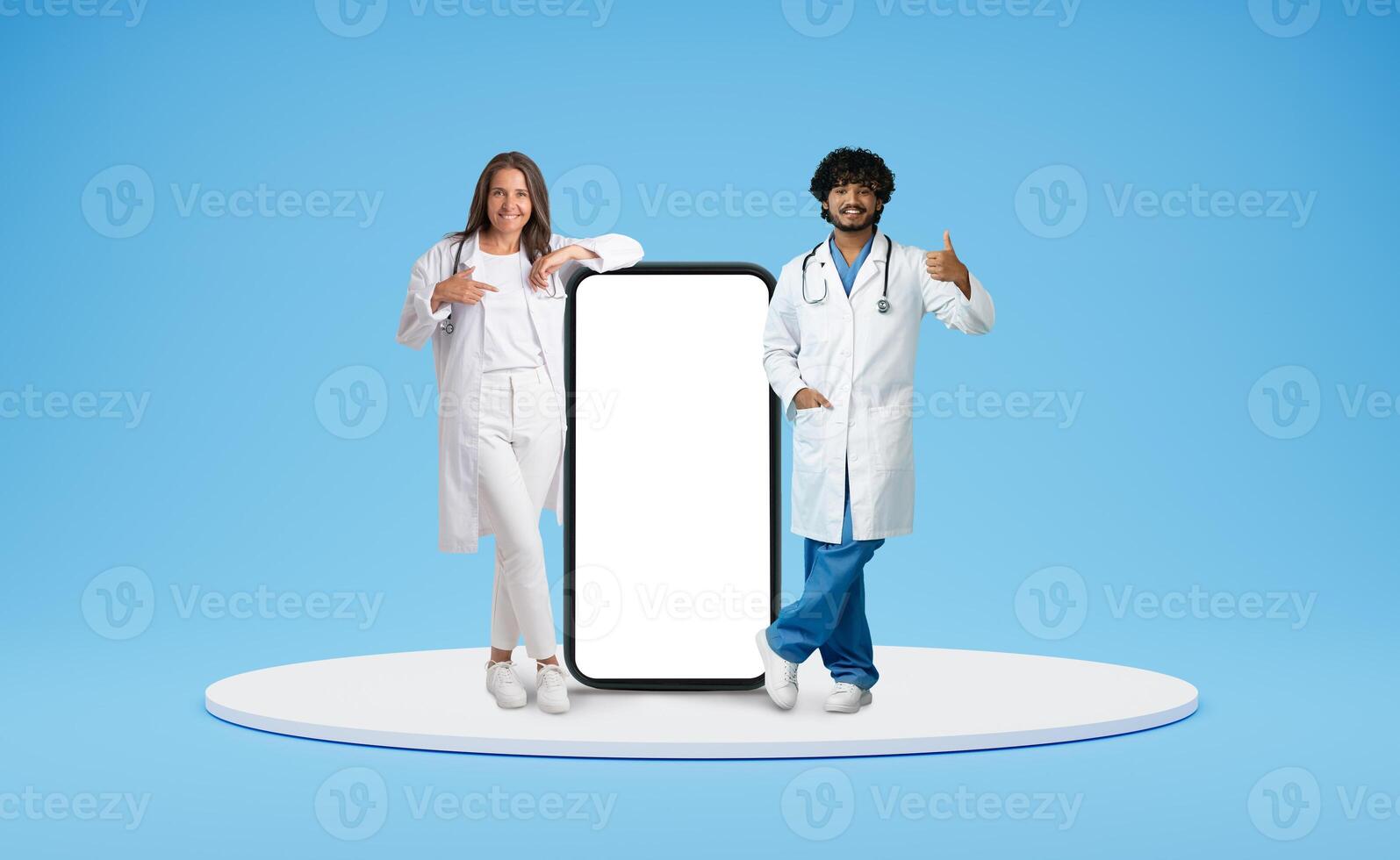 Telemedicine, virtual consultaion, ehealth. Cheerful multiracial doctors in medical workwear posing by huge phone with white blank screen, showing thumb up, isolated on blue background, mockup photo