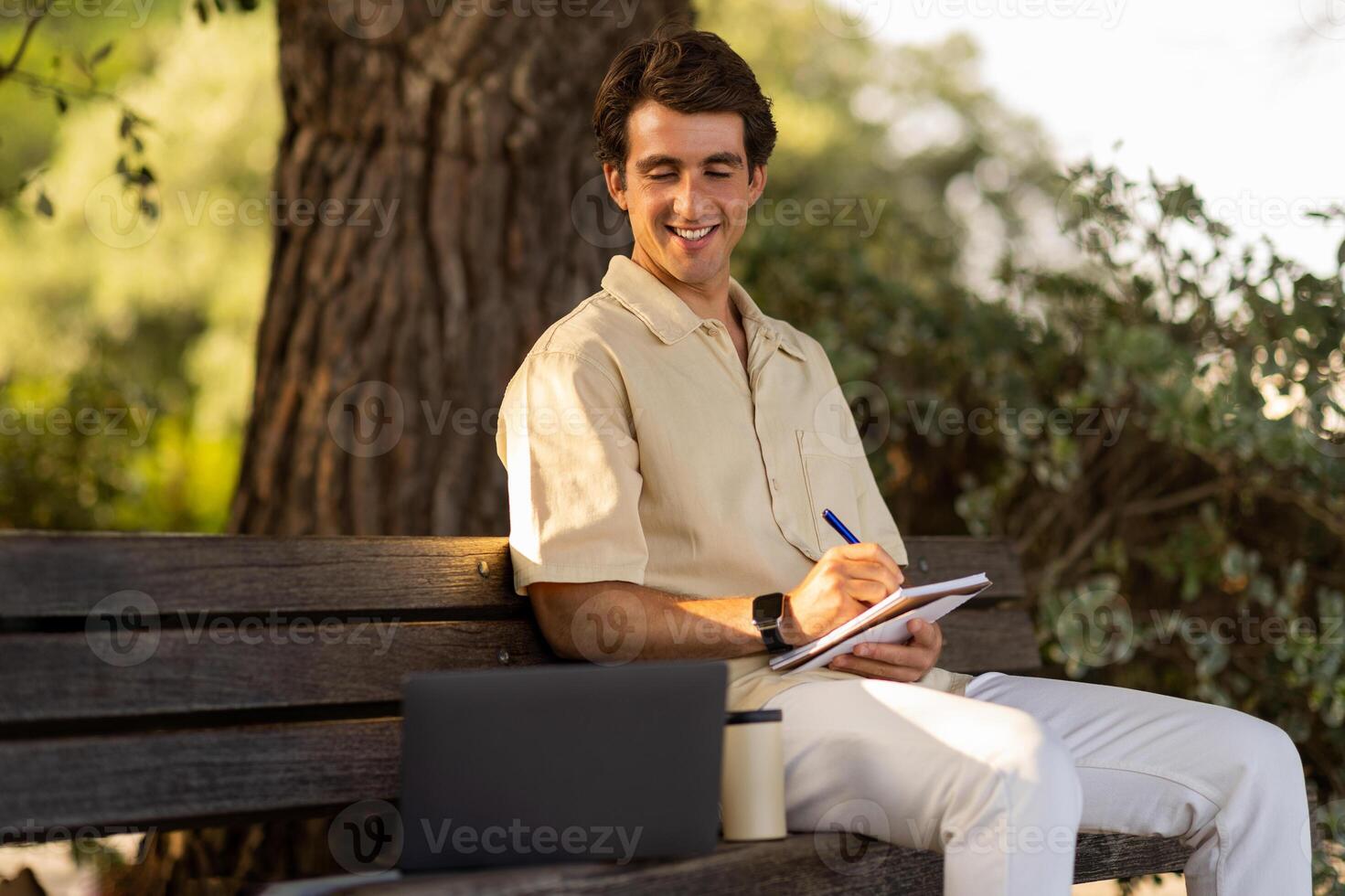 Cheerful young man attending webinar online course training while resting at park, guy sitting on bench at garden, looking at computer laptop screen, taking notes in notepad, learning new language photo