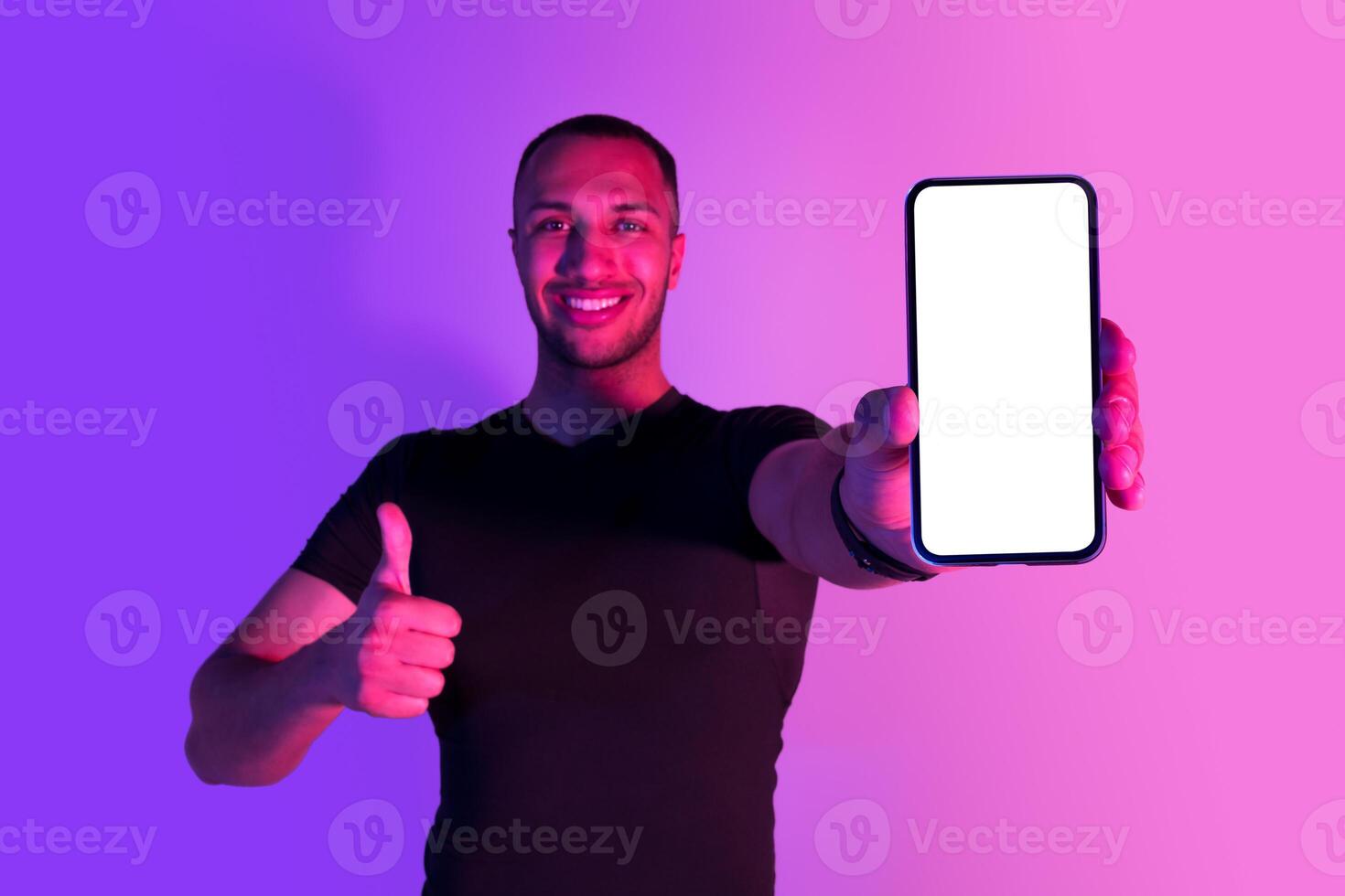 Happy black sportsman showing cellphone with empty screen and gesturing thumb up, smiling to camera, recommending workout and weight loss application, posing on purple neon background. Mockup photo