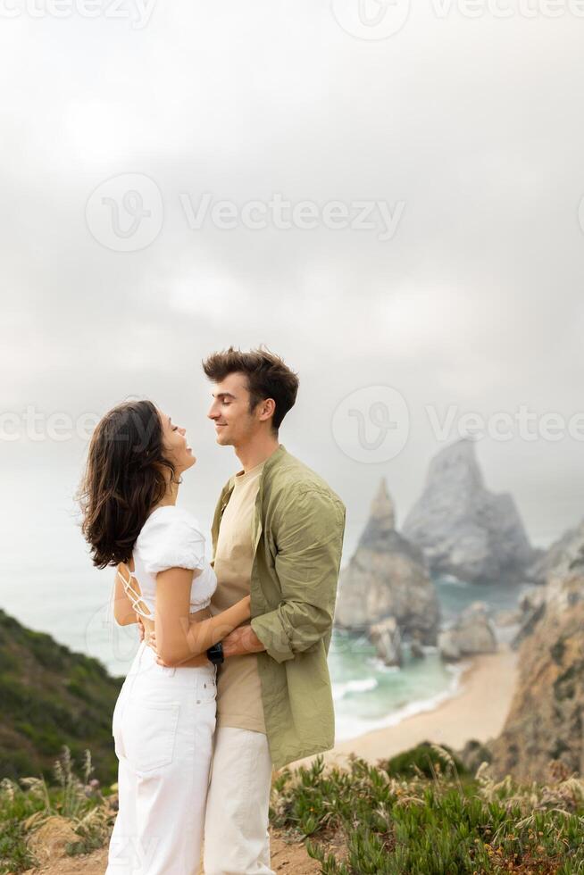 Young couple in love having romantic date, embracing and looking at each other, standing on coastline with beautiful view on ocean shore, vertical shot, copy space photo