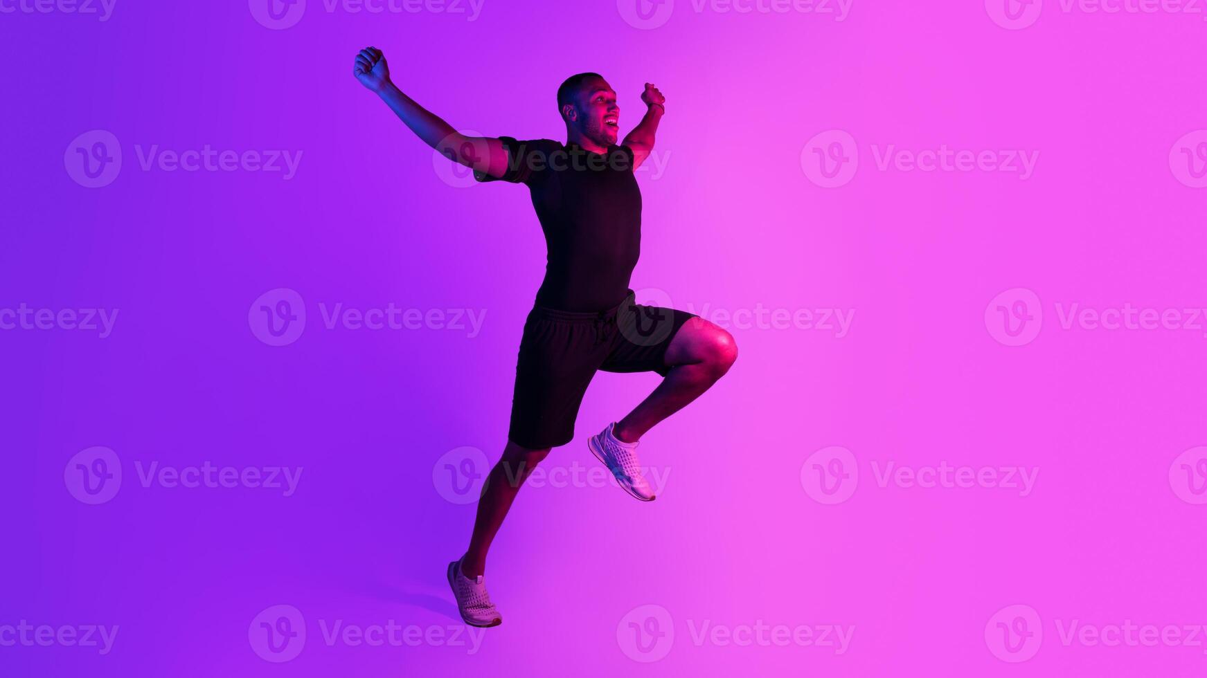 Sport victory, triumph. Emotional black runner man running raising hands and shaking fists in joyful gesture, celebrating win on purple neon background. Full length, free space for text. Panorama photo