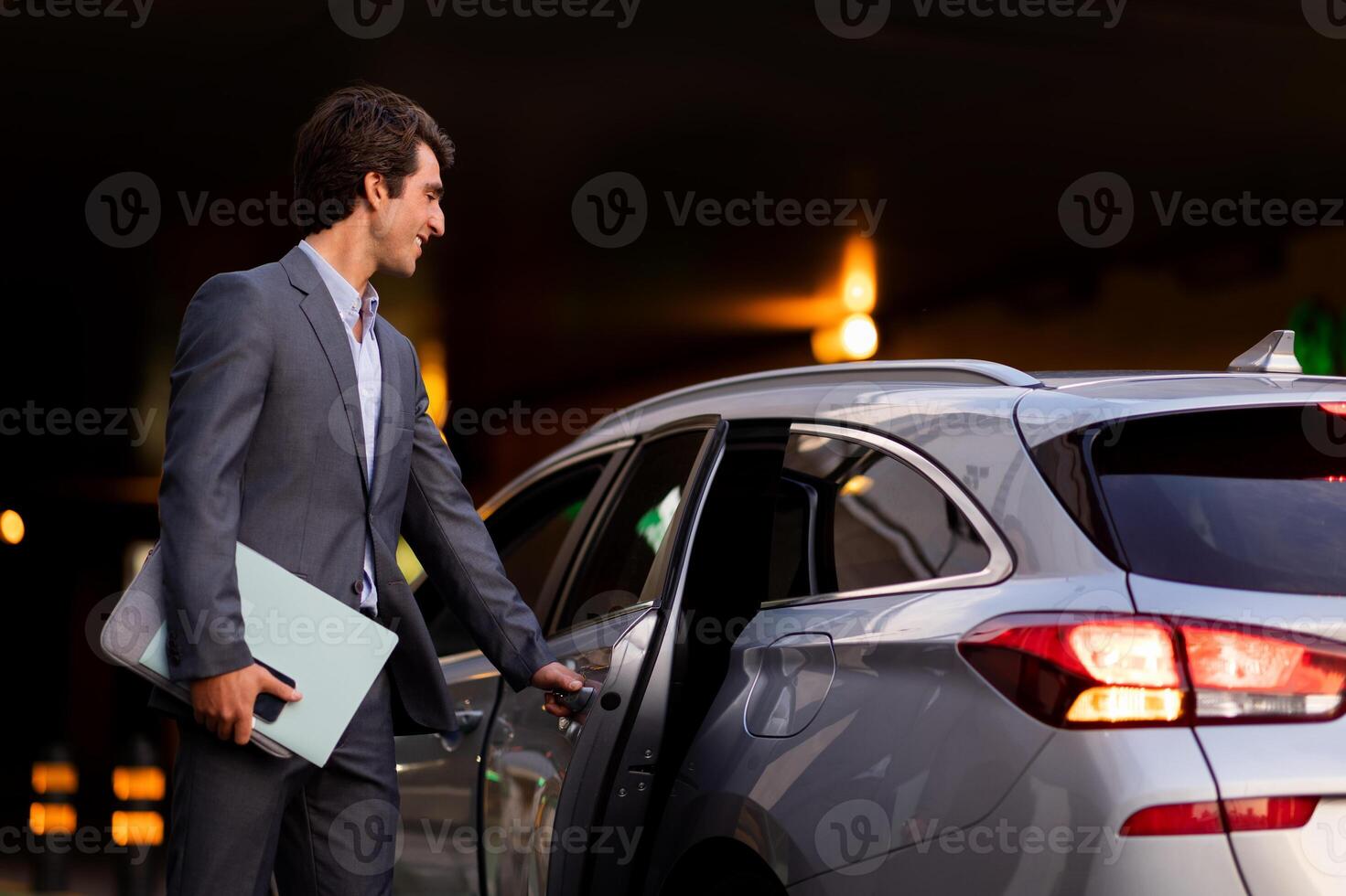 Handsome young businessman wearing expensive grey suit manager with laptop computer, phone and documents in his hand getting into luxury car in the evening, going home after busy day photo