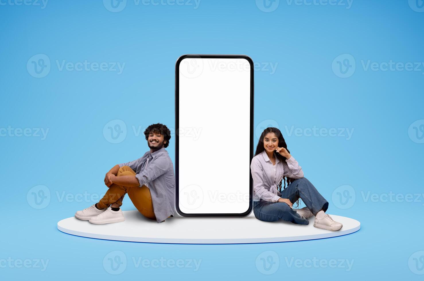 Cute cheerful smiling young eastern couple man and woman sitting on round platform by big phone with white blank screen, blue studio background, mockup blank space for mobile app, collage photo