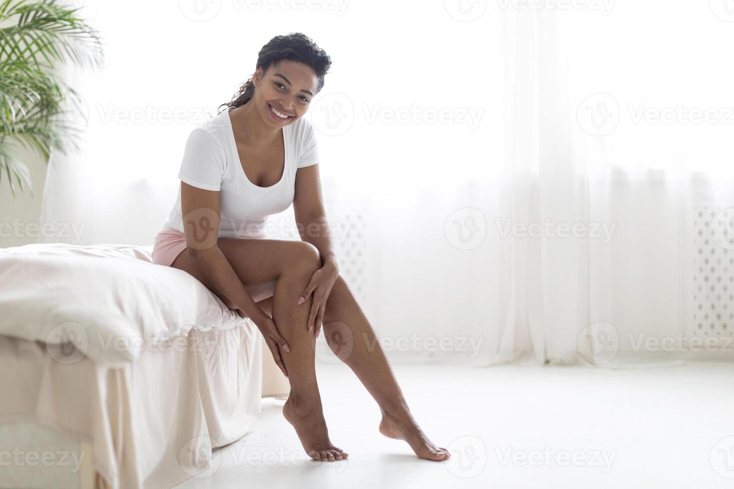 Beautiful young black woman touching her leg with silky skin after depilation, smiling african american female sitting on bed at home, attractive lady posing after domestic hair removal procedure photo