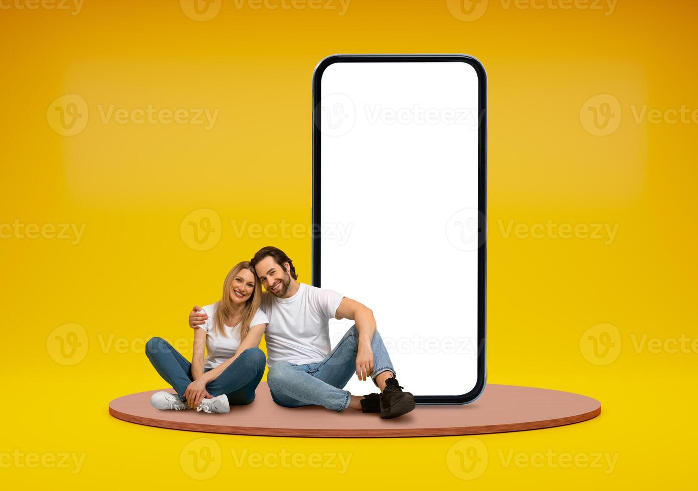 Happy cheerful beautiful young couple sitting on platform over yellow studio background, embracing next to huge phone with white blank screen, mockup for onlne offer summer deal, copy space photo