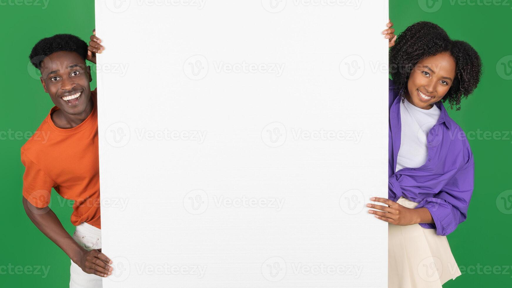 Smiling millennial african american couple in casual with big banner with blank space, isolated on green studio background, panorama. Recommendation together, people emotions, sale, ad and offer photo