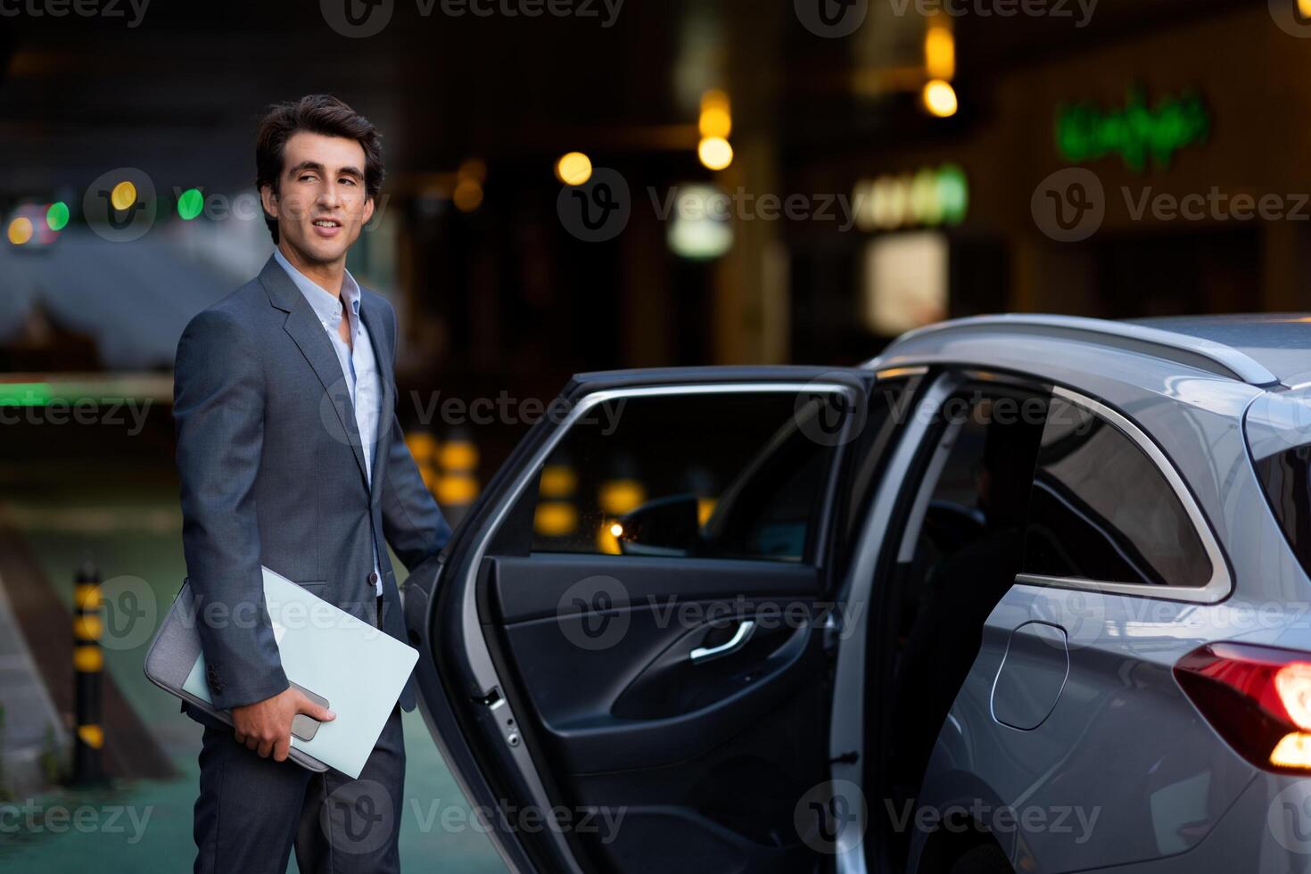 Attractive millennial man wearing expensive grey suit manager with laptop computer, phone and documents in his hand getting into luxury car in the evening, going home after working day photo
