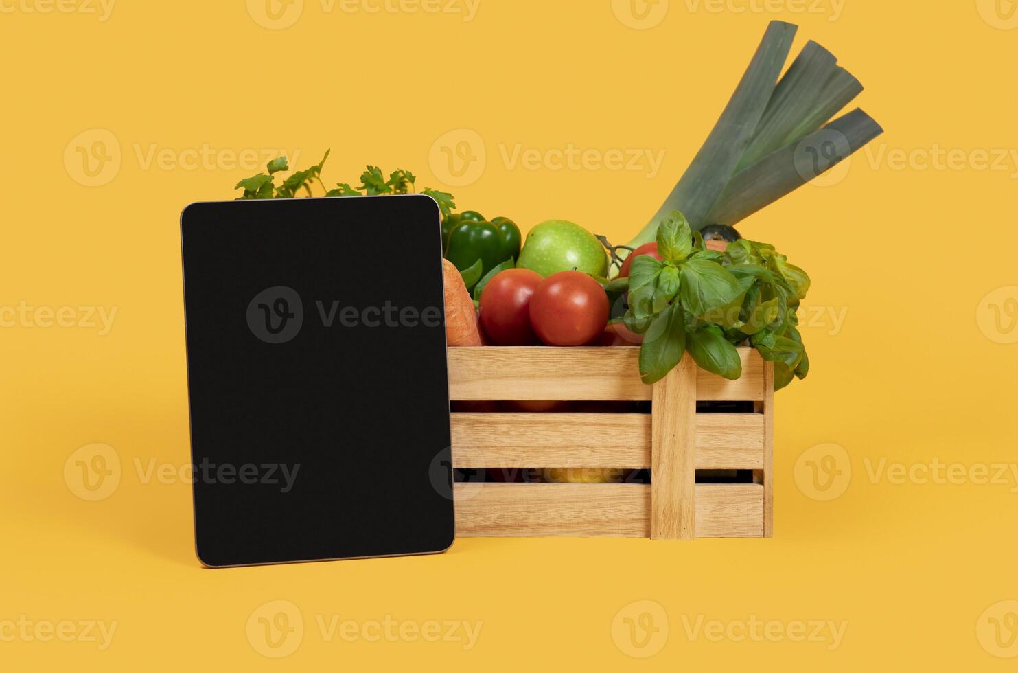 Digital Tablet With Blank Screen Standing Near Wooden Box With Fresh Vegetables And Fruits Over Yellow Studio Background. Mockup For Agriculture Website Advertisement Or Online Grocery Shopping photo