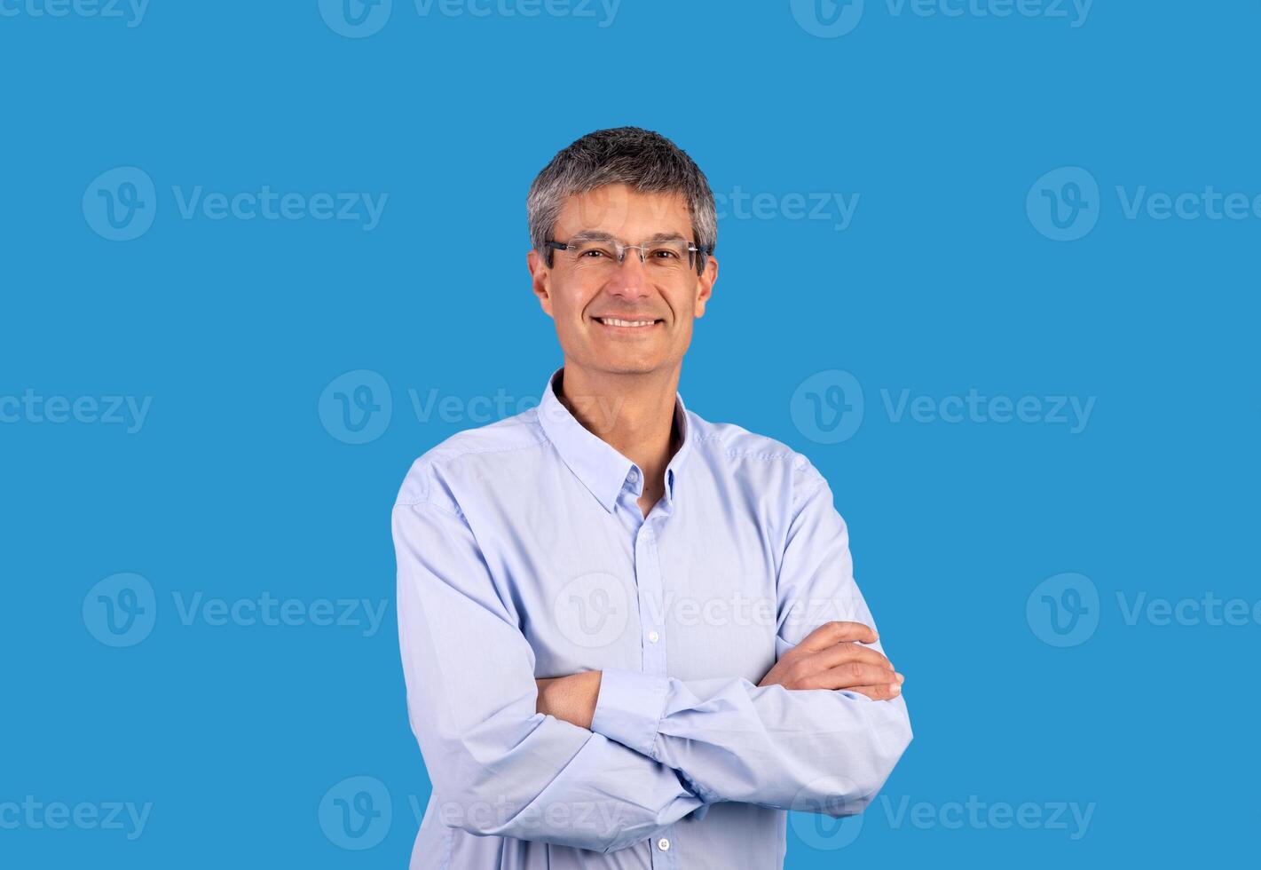 Confident business person. Happy mature businessman in shirt posing and crossing hands with expression of confidence, smiling looking at camera standing over blue background, studio shot photo