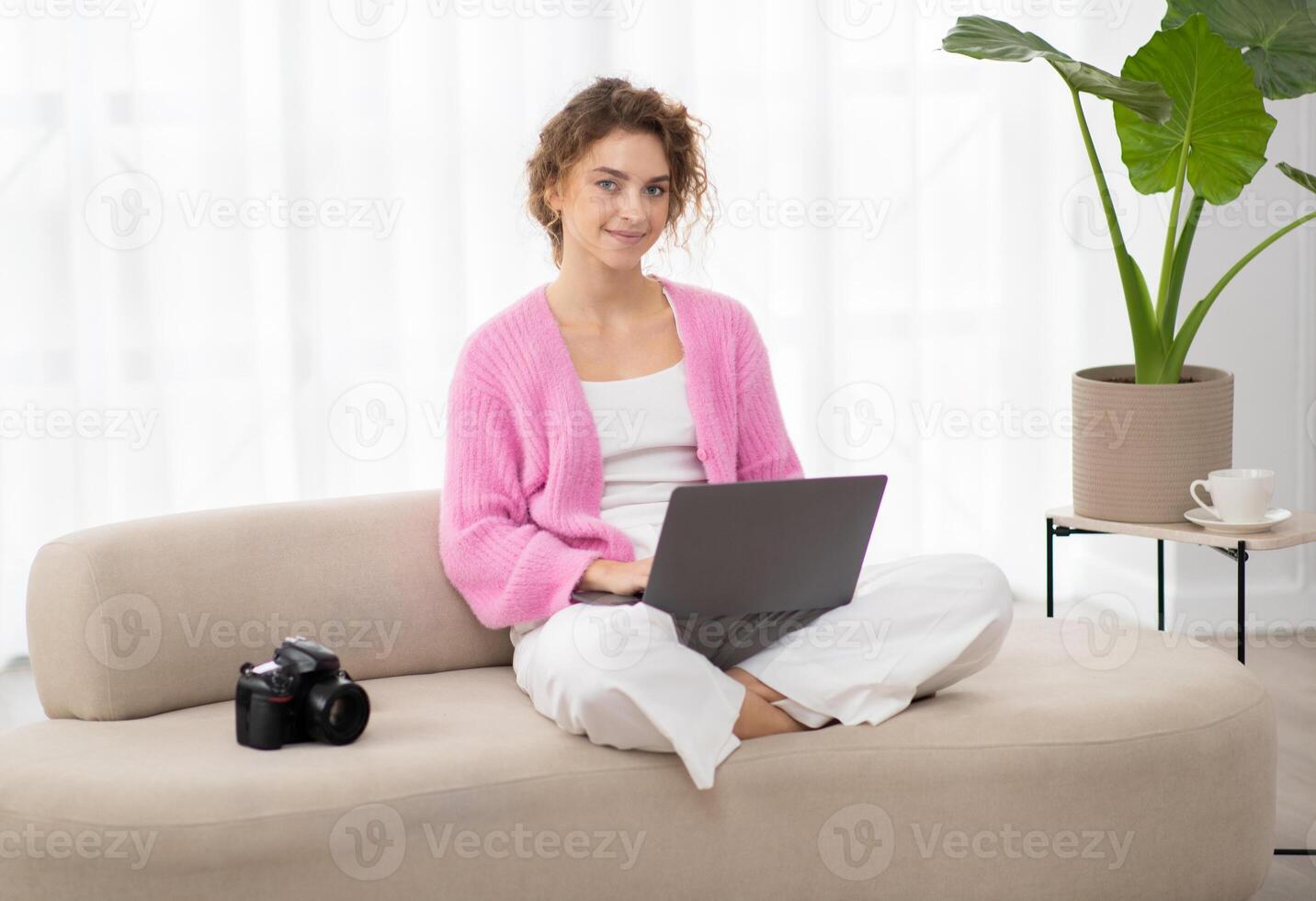 Portrait Of Smiling Young Woman With Laptop Sitting On Couch Next To Photocamera, Beautiful Millennial Freelance Photographer Lady Using Computer At Home For Editing Photos, Copy Space photo