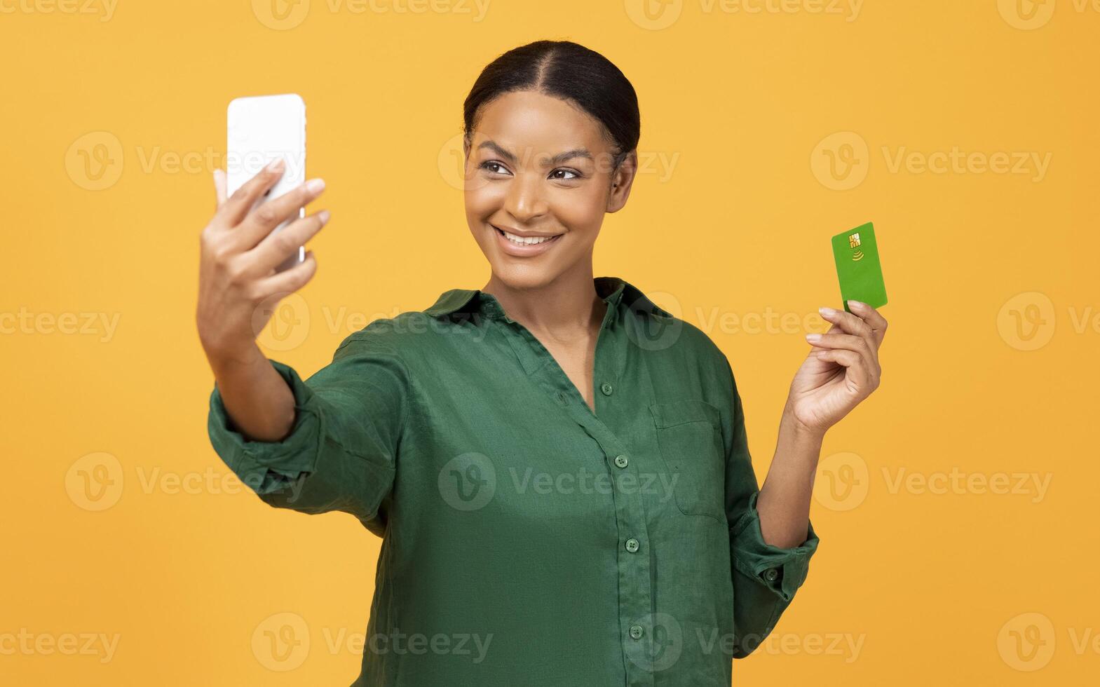 Mobile Shopping App. Happy African American Lady Holding Phone And Credit Card, Making Selfie Posing With Bankcard And Using Financial App Standing Over Yellow Studio Background photo