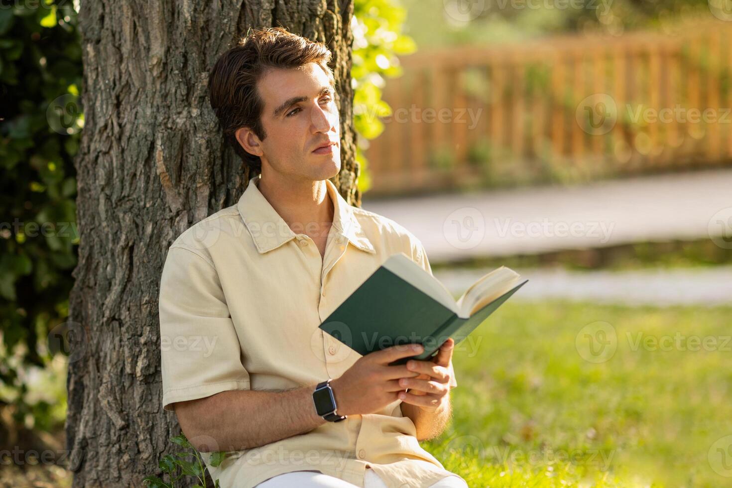 Handsome relaxed young guy in beige shirt sitting under tree at public park or garden, reading book, looking copy space, thinking about story. Relaxation, stress relief, weekend, rest concept photo