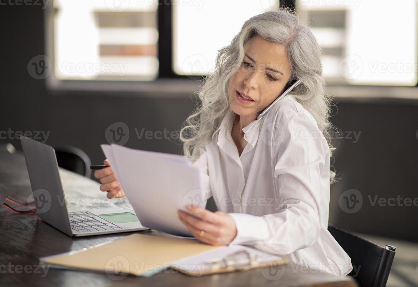 Business Communication. Serious Middle Aged Businesswoman Talking On Phone Holding Papers, Discussing Documents Issues And Concerns During Telephone Conversation In Modern Office photo