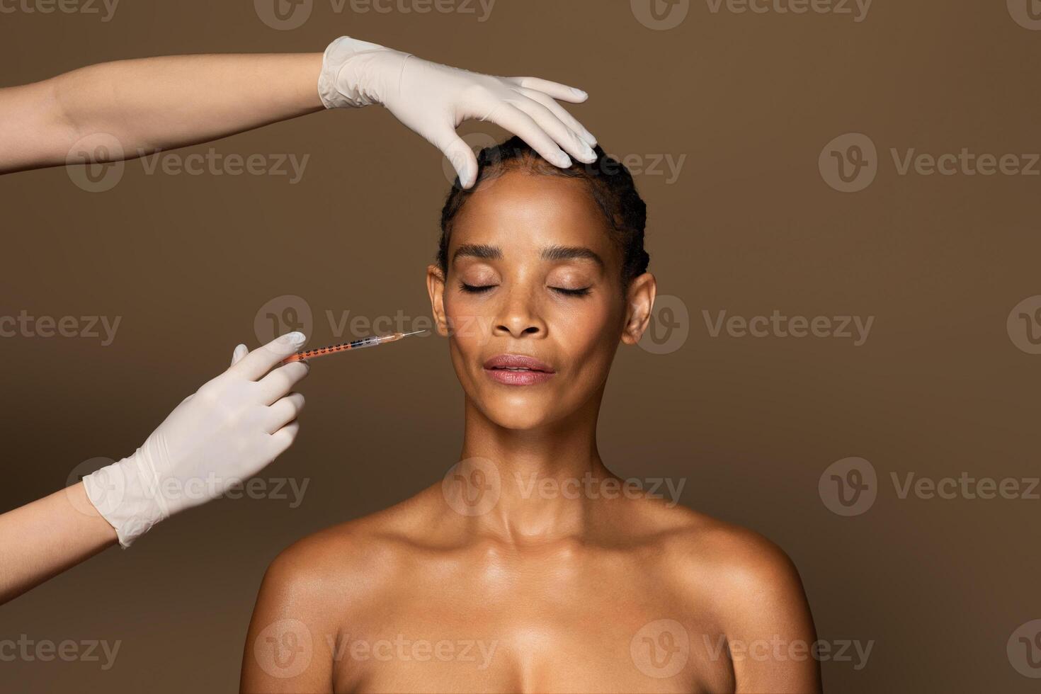 Beautiful african american middle aged woman getting facial injection in cheekbones zone, standing over brown studio background. Cosmetic beauty injections concept photo