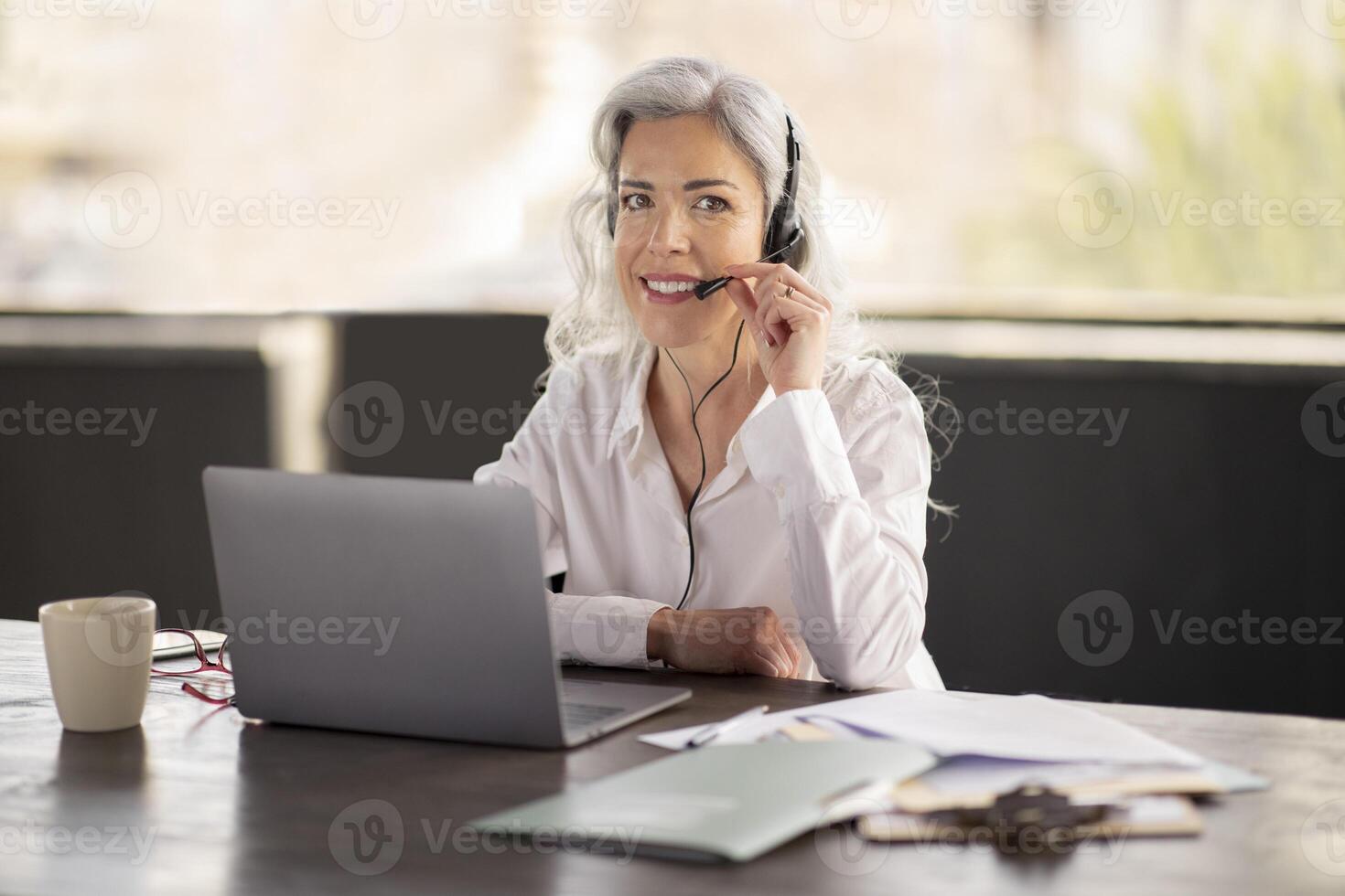 Online Helpline. Businesswoman Calling On Laptop Wearing Headset, Smiling At Camera While Communicating With Remote Client, Offering Business Expert Support Sitting In Modern Office Interior photo