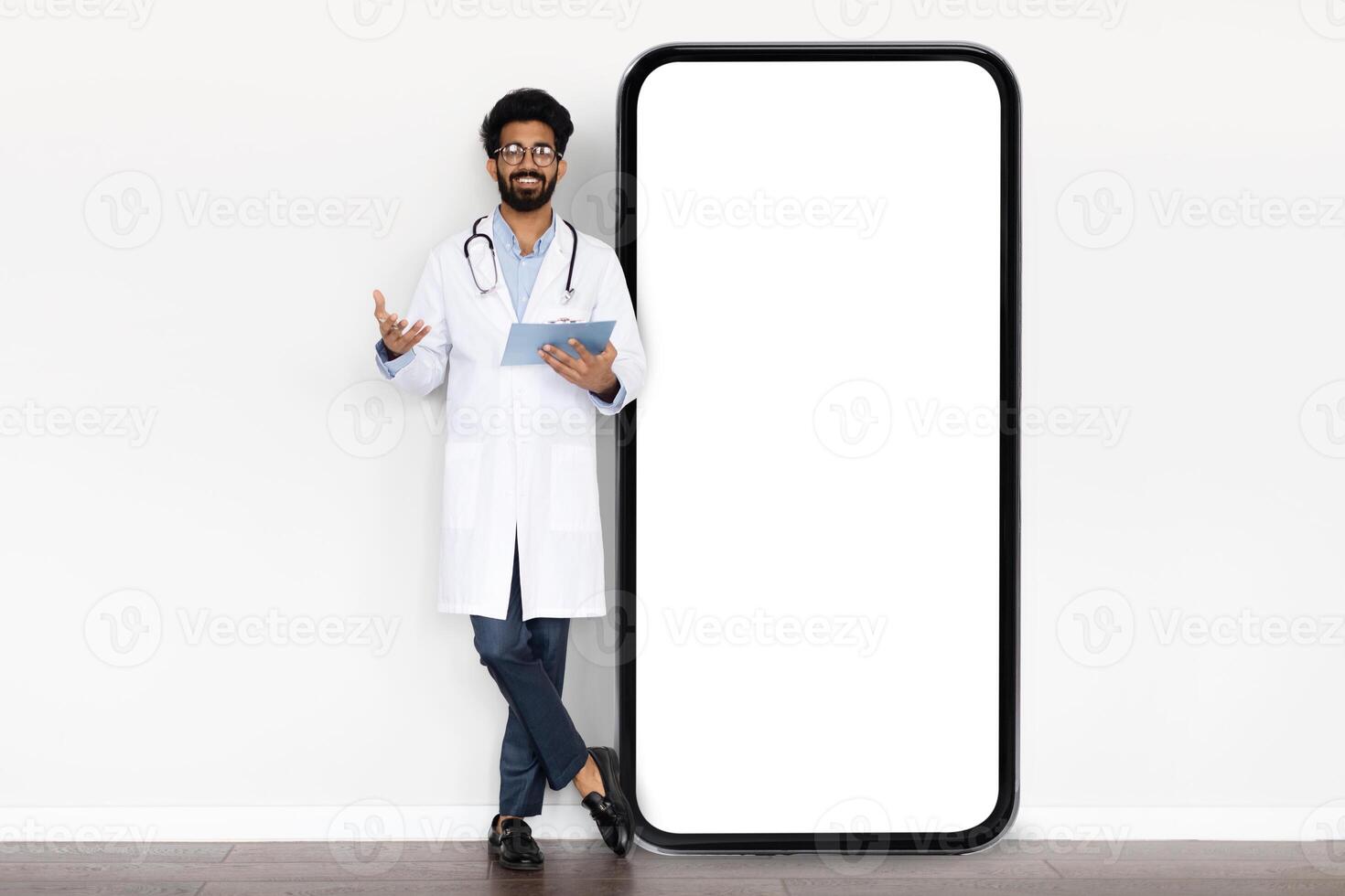 Telehealth app. Positive handsome millennial eastern doctor holding medical chart wearing white medical coat standing by huge smartphone with white empty screen mockup, isolated on white background photo