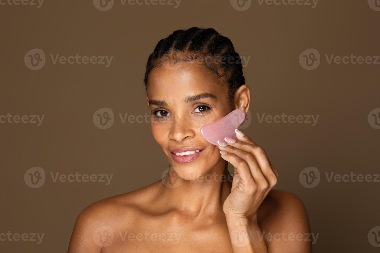 Sensual half-naked black middle aged woman using pink gua sha stone, massaging face with scraper, smiling at camera isolated on brown background, free space photo