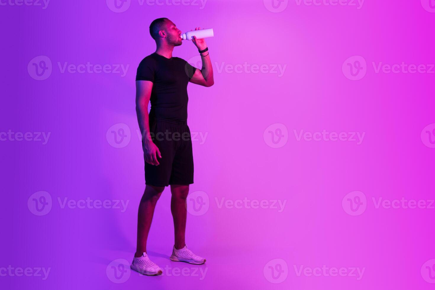 Young black sportsman in sportswear drinking water on purple studio background, posing with fitness bottle, standing near free space. Full length of sporty guy staying hydrated after workout photo