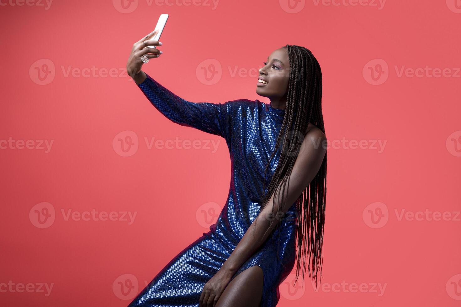 Smiling African American Woman In Sequin Dress Taking Selfie On Smartphone While Sitting On Chair Over Red Studio Background, Stylish Young Black Female Making Self-Portrait, Posing At Mobile Camera photo