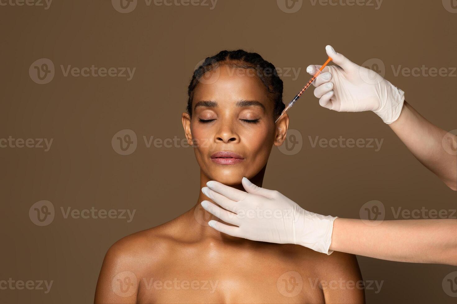 Cosmetic beauty injections concept. Attractive black middle aged woman getting facial injection in cheekbones zone, standing over brown studio background, free space photo