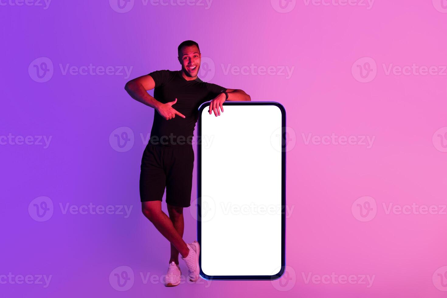 Young black fitness guy standing near huge smartphone with blank screen, pointing finger at device demonstrating mobile application for online workout over purple studio background, mockup photo