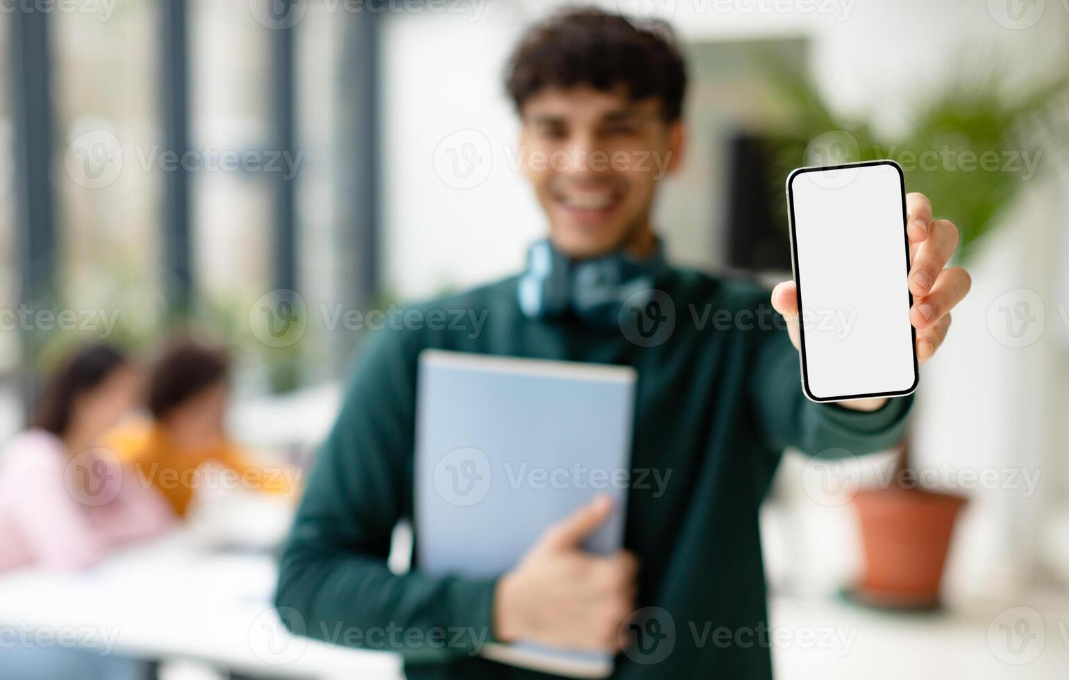 Educational advertisement. Happy male student showing smartphone with white blank screen, mockup, selective focus. Guy standing in coworking space with classmates on background photo