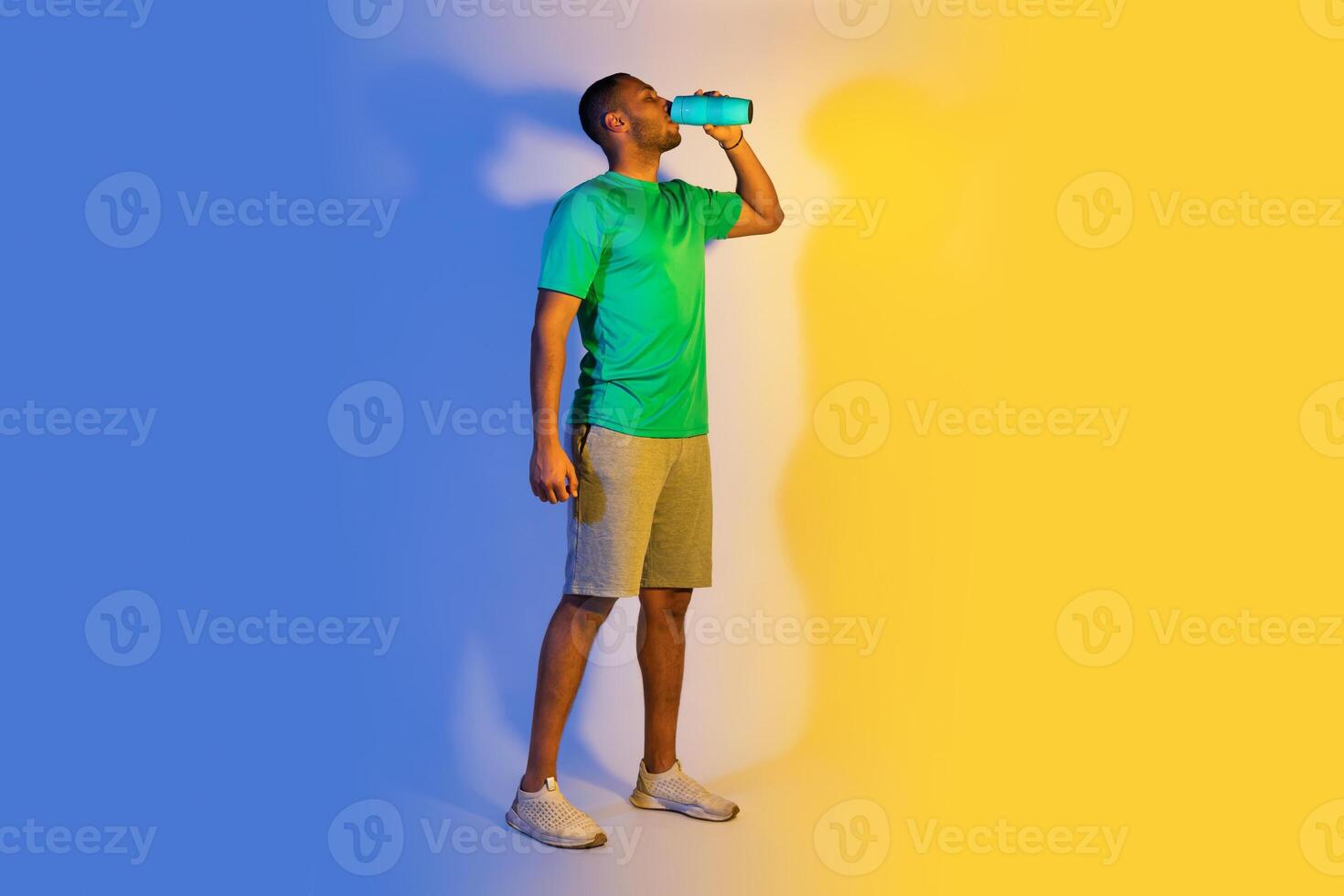 Sport, Hydration And Weight Loss. Full Length Of Young Black Sportsman Drinking Water From Bottle, Hydrating During Workout Training Standing Over Blue And Yellow Studio Background, Free Space photo